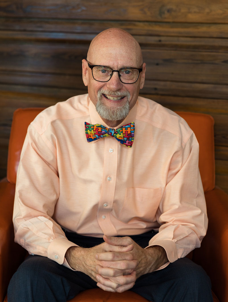 Mr. Bob Barrows wearing an orange shirt and bow tie sitting with hands together in front of him
